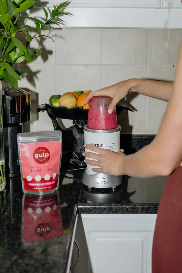 A hand fitting a blender cup of pink smoothie onto its base, next to a Gulp Beet Bliss pouch